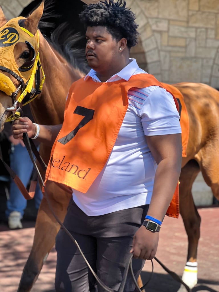 Man in orange racing silks holding reins of brown horse with yellow racing bib, standing outdoors near brick wall